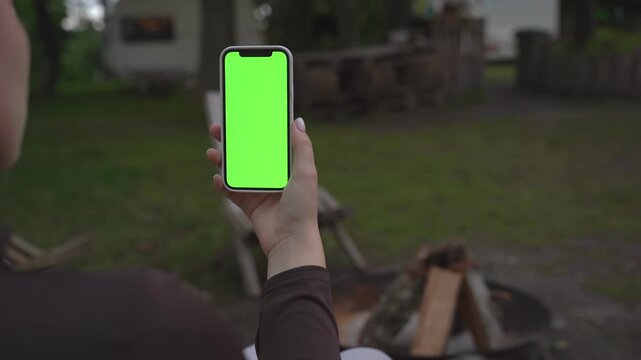 Person holds phone with green screen outdoors. Focus on hand and device. Background shows backyard and firewood. Ideal for app ads, social media, or digital content