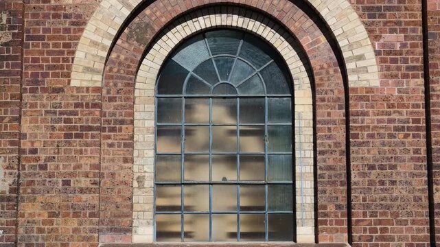 Beautiful arched factory window and surrounding brickwork from nineteenth century industrial site with interesting texture under beautiful sunlight. Camera tilts up from window base to reveal arch.
