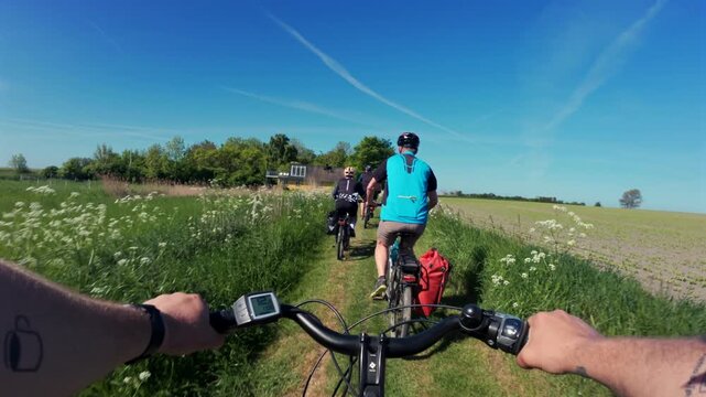 First-person chest-mounted view riding an electric bike behind a group of cyclists on a farm-field section of the EuroVelo 10 route in Denmark on a sunny summer day
