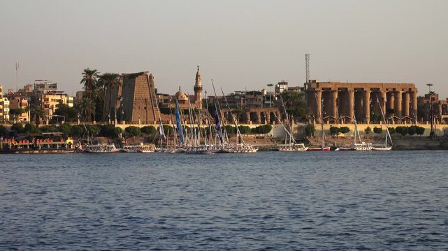 View of traditional felucca boats sailing on the Nile River with the historic Luxor Temple in the background in Luxor Governorate, Egypt.