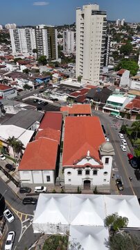 Imagem de drone da igreja no Socorro, em Mogi das Cruzes, S&atilde;o Paulo, com destaque para a arquitetura religiosa, o telhado de cer&acirc;mica vermelha e o entorno urbano com ruas, casas e edif&iacute;cios