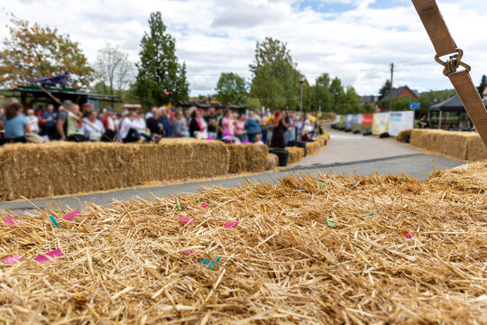 Hay bales line roadside race course while spectators gather behind safety barriers during outdoor soapbox racing event. Straw foreground and blurred crowd create festive rural competition atmosphere