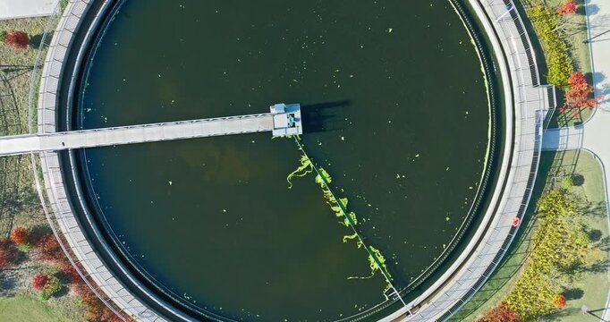 An aerial top-down view of a circular wastewater treatment tank filled with dark water, green algae, and a metal bridge under bright sunlight.