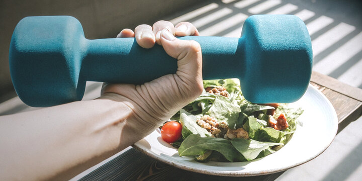 Exercise and healthy eating lifestyle with a hand holding a dumbbell above a bowl of fresh vegetable salad