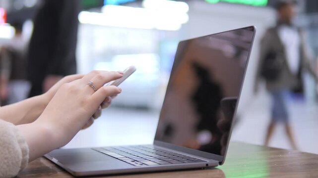 Close-up of hands using smartphone near open laptop. Blurred background with people moving. Modern indoor setting with wooden table. Focus on interaction between mobile and laptop devices