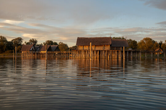 Reconstructed prehistoric stilt houses glow in the golden light of sunset over the calm waters of Lake Constance.