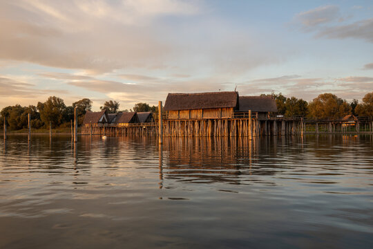 Reconstructed prehistoric stilt houses glow in the golden light of sunset over the calm waters of Lake Constance.