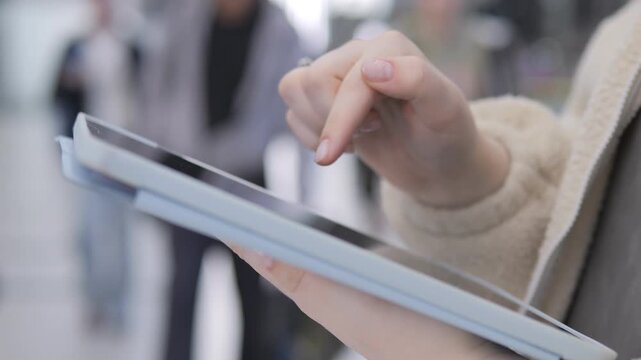 Person holds tablet, finger taps screen. Blurry background suggests public space. Close-up on hand and device only. Neutral tones, soft lighting. No animals or plants visible