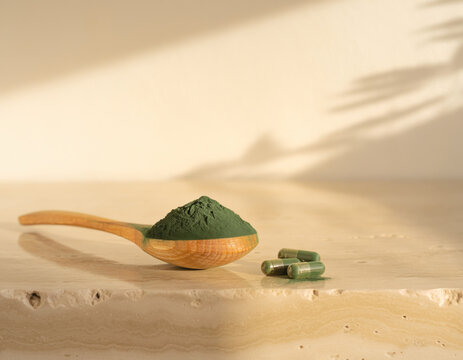 Green spirulina powder in a simple wooden spoon, accompanied by several natural supplement capsules, rests on a light stone surface with gentle shadows, healthy lifestyle and natural nutrition