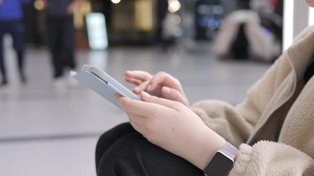Person uses phone while sitting in public space. Hands hold device close to body. Background shows blurred people and lights. Wears beige coat and black pants. Wristwatch visible on left arm