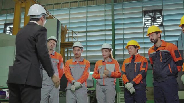 A manager in a suit and hard hat briefs a diverse team of factory workers, assigning tasks and responsibilities while standing together inside an industrial manufacturing plant.