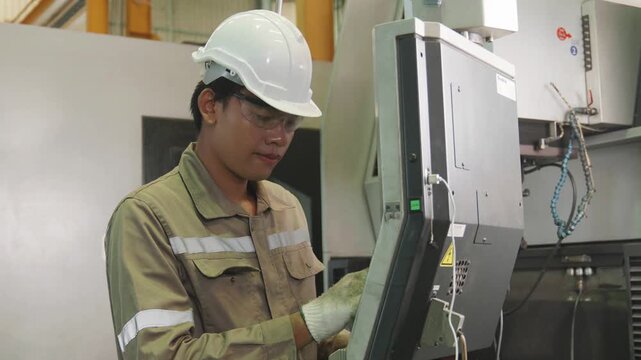 A male technician in a hard hat and safety glasses operates a CNC control panel, adjusting settings on industrial machinery inside a modern manufacturing factory workshop.