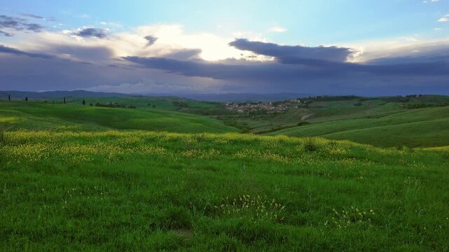 View of rolling green hills and yellow wildflowers under a dramatic cloudy sky in the countryside of Tuscany, Italy.