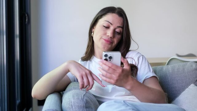 Beautiful young woman with long brown hair relaxing on a comfortable couch in her living room, smiling while scrolling and tapping on her smartphone's touchscreen for online shopping