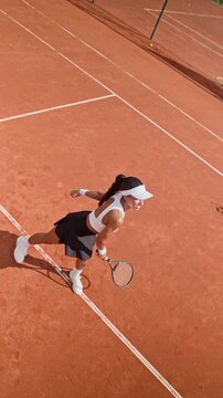 Vertical video of young adult Biracial woman serving tennis ball on clay court in slow motion from high angle. Ideal for mobile sports apparel advertising, fitness apps and athletic campaigns