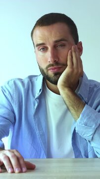Young handsome bearded man sitting at a desk and looking at camera with a bored, pensive, and uninterested expression, feeling lonely and tired of waiting isolated on a white background