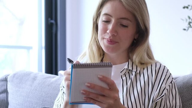 Beautiful thoughtful woman sitting on a cozy sofa, writing down her plans, goals, or a to-do list in a small spiral notebook, feeling inspired and focused on her creative process