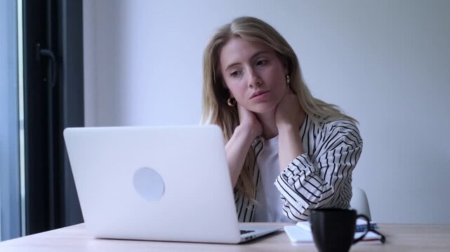 Exhausted young businesswoman feeling neck pain and stiffness from sitting at her computer for too long, experiencing fatigue and discomfort due to overworking in her modern office space