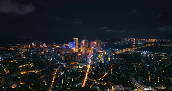 Aerial view of the Macau cityscape illuminated at night, featuring glowing skyscrapers, city streets, bridges over water, and dark night sky.