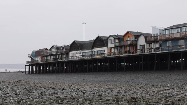 View of buildings on stilts along the Homer Spit with wooden pilings and a pebble beach under an overcast sky in Homer, Alaska, United States.