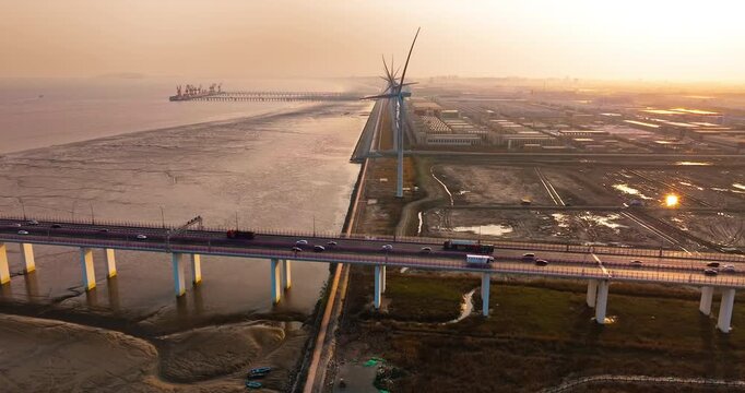 Aerial view of an elevated highway bridge with vehicles near wind turbines and coastal mudflats, lit by warm golden sunset light.