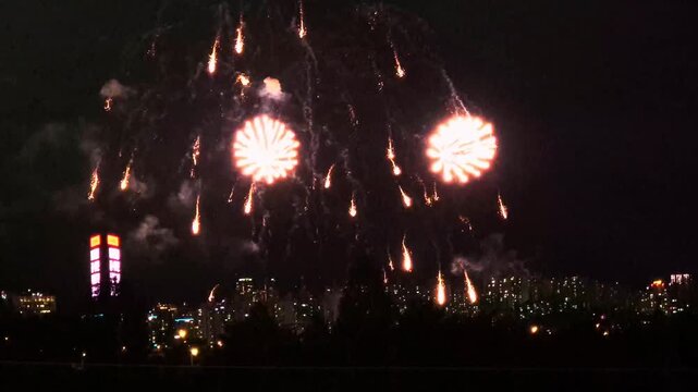 Individual fireworks shells bursting one by one in orange, white and red over the Seoul city skyline at night, above the Han River in South Korea