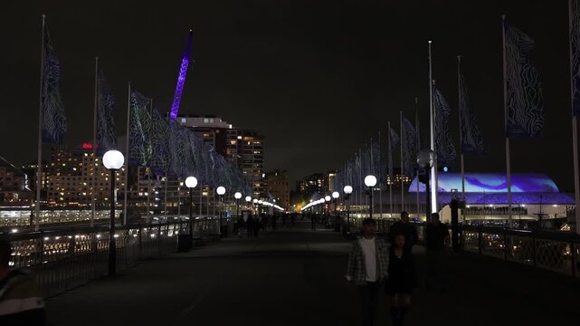 View of people walking along the illuminated Pyrmont Bridge at night with city lights and decorative flags in Sydney, New South Wales, Australia.