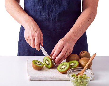 Femme en tablier bleu d&eacute;coupant des kiwis frais sur une planche &agrave; d&eacute;couper pour pr&eacute;parer une salade de fruits dans un bol en verre