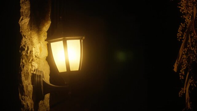 Light from a wall lamp shines brightly in the night, illuminating the dark surroundings near a stone wall
