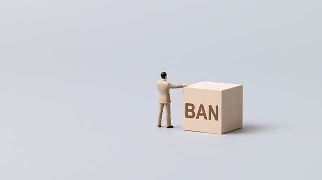 A miniature businessman figure stands beside a wooden block displaying the word ban on a clean grey background to represent business policy