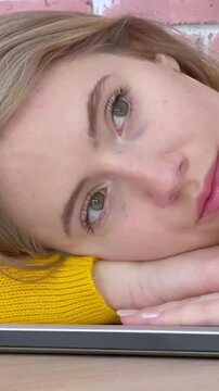 Beautiful thoughtful young woman with blonde hair resting her head on a closed laptop, feeling bored and unmotivated while procrastinating from work or study in a modern loft space