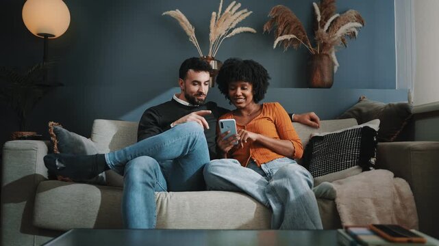 Multi racial couple using mobile phone sitting together on sofa at cozy home