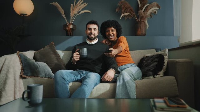 African American and Hispanic caucasian couple sitting at home on sofa watching tv movie online streaming platform show 