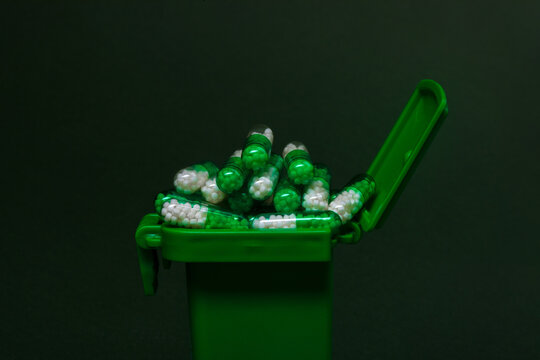 Overflowing green medical waste bin filled with white and green capsules against a dark background. Concept for healthcare, pharmacy, and waste management.