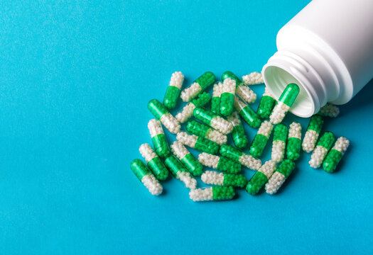 White plastic bottle with green and white capsules spilled on blue background, representing healthcare, medicine, and pharmaceuticals.