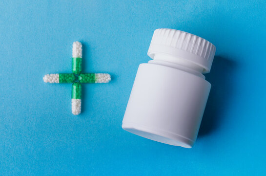 White medicine bottle and green capsule cross on blue background. Concept for healthcare, pharmacy, or medication.