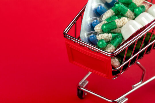 Mini shopping cart filled with assorted capsules and tablets on a vibrant red background, symbolizing healthcare, pharmacy, and medical purchases.