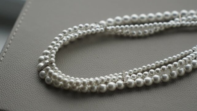 Close up of a multi strand pearl necklace laid on a grey faux leather box. The composition emphasizes the pearly sheen bead spacing and textured surface of the box. Soft diffused light and shallow dep