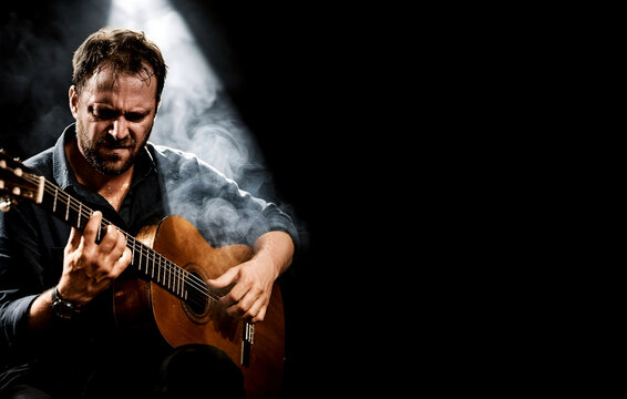 Dramatic Acoustic Guitarist With Copy Space. Male musician plays acoustic guitar under a spotlight with smoke and black background, ideal for music ads, posters, banners and editorial use.