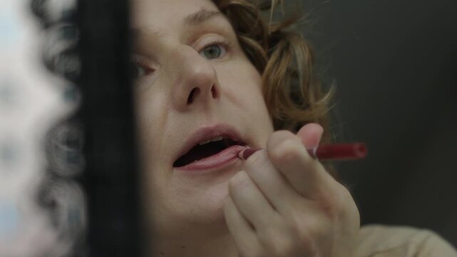 Close up of a woman applying a lip liner pencil while looking into a mirror. The mirror reflection shows her face and hand as she carefully draws the lip line. Soft natural light and shallow depth of 