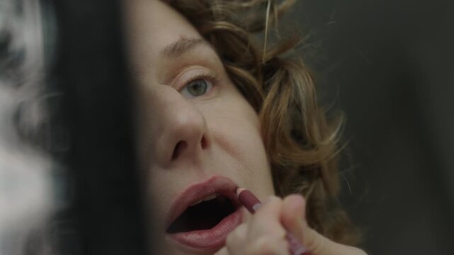 Close up of a woman applying a lip liner pencil while looking into a mirror. The mirror reflection shows her face and hand as she carefully draws the lip line. Soft natural light and shallow depth of 