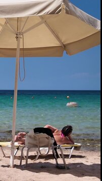 Young woman relaxes on a sun lounger on a sunny beach by the crystal clear sea., a beachgoer resting on a deckchair with a cigarette in his hand. Vertical view.