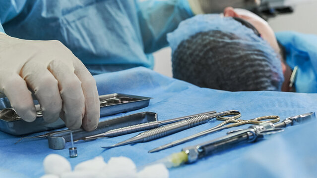 Blurred close-up of surgical instruments arranged on sterile drape, with faceless dentist performing dental implant procedure in background, creating professional oral surgery setting.
