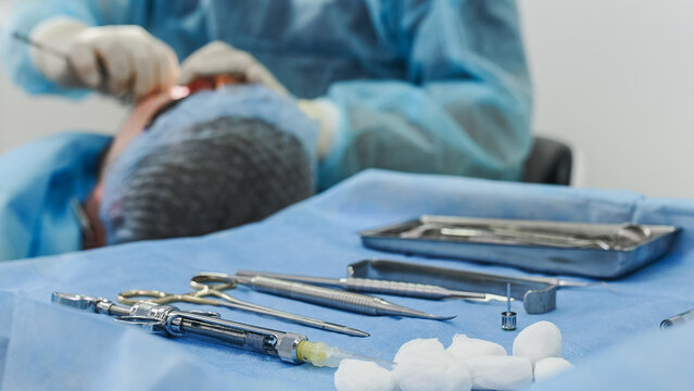 blurred close-up of surgical instruments laid out on a sterile drape on a table. A surgeon, faceless and unrecognizable, is in the background. An orthopedic dentist performs a dental implant.