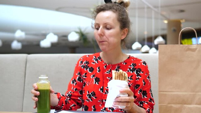 Woman in red floral top bites wrapped sandwich and sips green juice at cafe booth, young adult woman with concentrated expression takes relaxed snack break beside paper bag, eating and drinking.