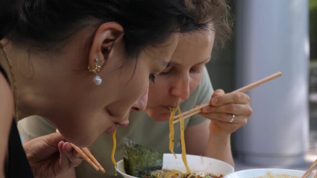 Two women eat noodles with chopsticks during casual outdoor dining, one focused woman leaning in to taste bowl of steaming noodles. Japanese style meal service adds relaxed social lunch moment.
