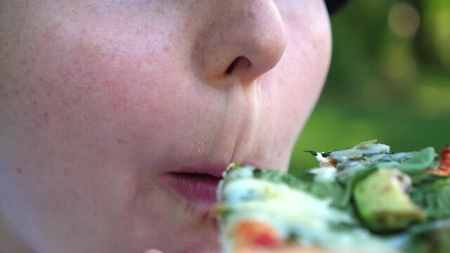 Young adult woman eats pizza in outdoor setting with calm, focused expression and natural style. Woman chews another bite, holding slice near mouth as green background stays softly out of focus.