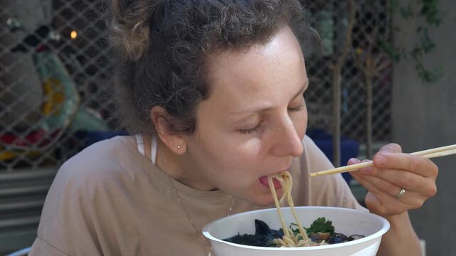 Young adult woman eats noodles with chopsticks, intent and concentrated, in casual outdoor style. Woman holds white bowl close and slurps strands, taking small bites at outdoor table.