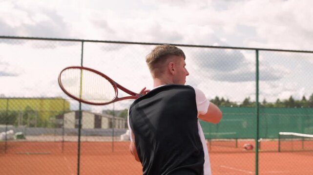Caucasian young adult man practicing forehand stroke on clay tennis court in slow motion, tilt up shot. Perfect for sports training videos, athletic brand commercials and tennis club marketing