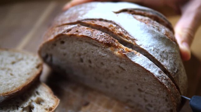 Bread loaf is being cut into even slices with serrated knife, rustic style and steady hands showing precise bread preparation on wooden board, producing thick slices with textured crust and crumb.